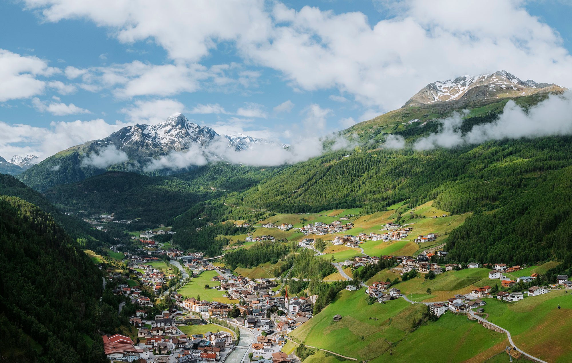 A picturesque mountain village lies in a green valley, surrounded by forests and snow-capped peaks, beneath a partly cloudy sky.