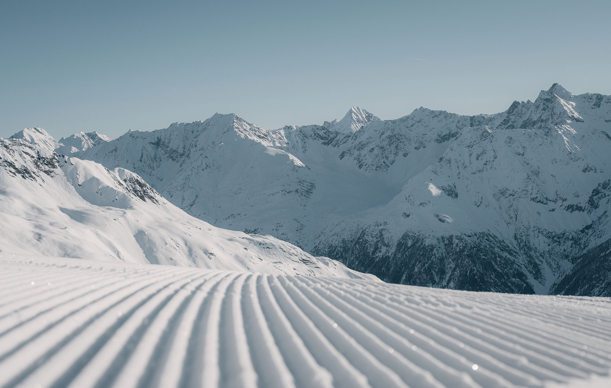 A groomed ski slope with parallel tracks in the snow, set against a backdrop of high, snow-capped mountains under a clear sky.