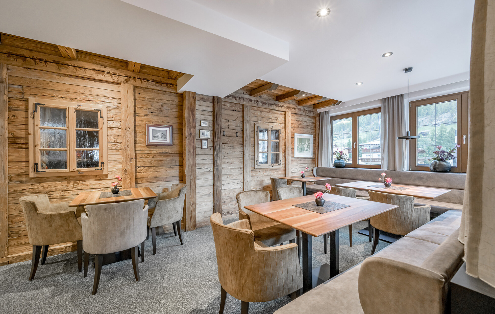 Cozy bar area with wood-paneled walls, light brown chairs, wooden tables, window-side bench seating and small flower vases on every table. Sunlight streams in through large windows offering a view outside.