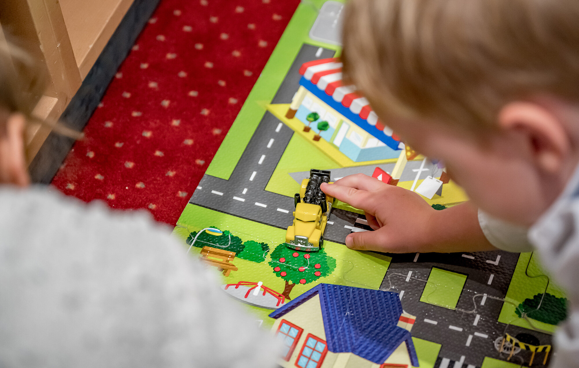Two children are playing on a colorful play mat featuring roads and buildings, with one of the children pushing a small yellow toy truck along the road.