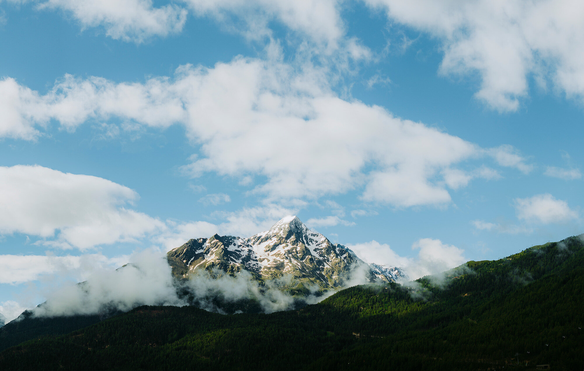 A snow-capped mountain peak rises above green, wooded hills, while scattered clouds are in the brilliant blue sky.