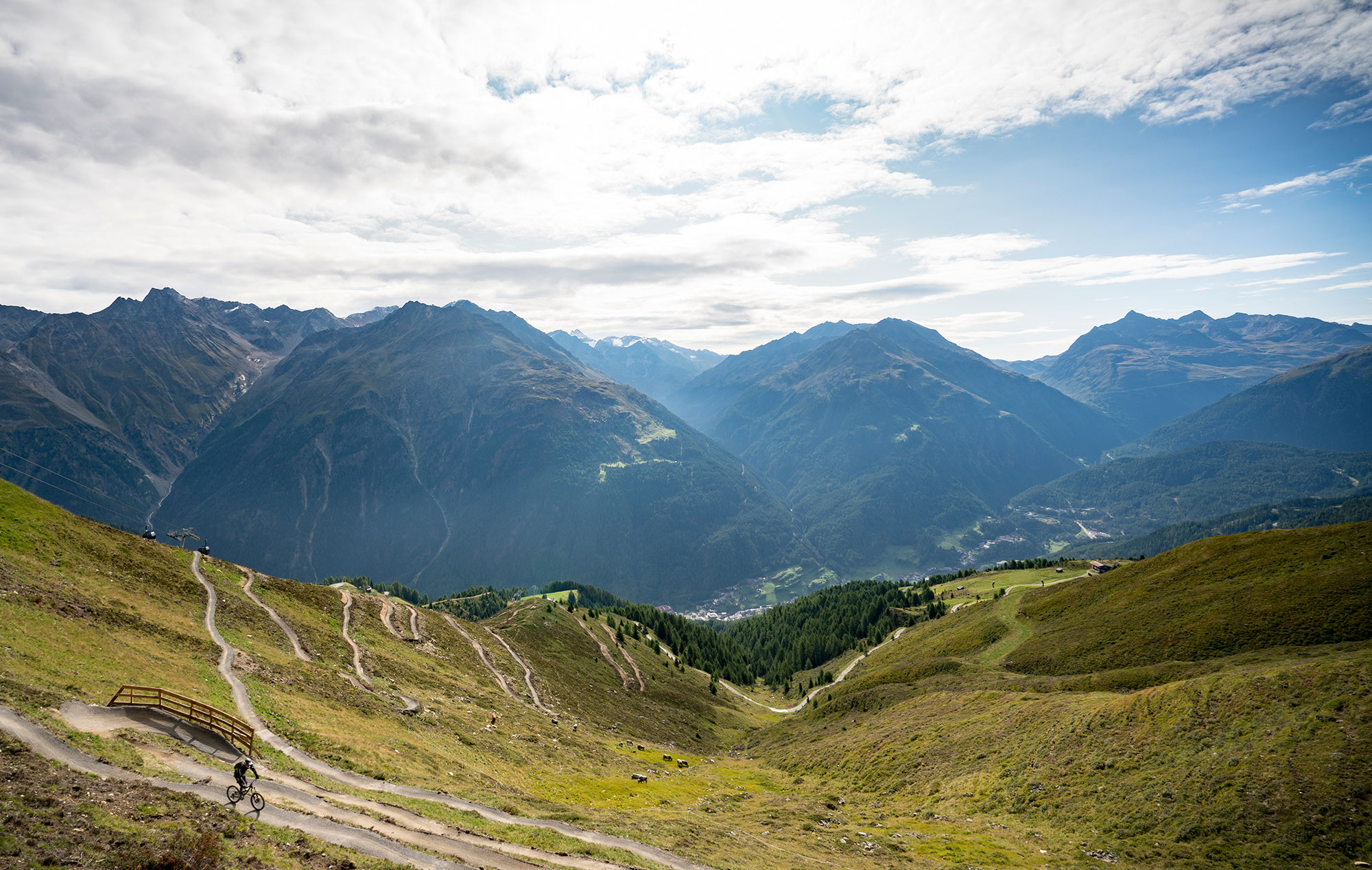 A winding mountain path leads down a grassy slope towards a valley, surrounded by high, rugged peaks under a partly cloudy sky. In the bottom left corner, a cyclist rides along the path.