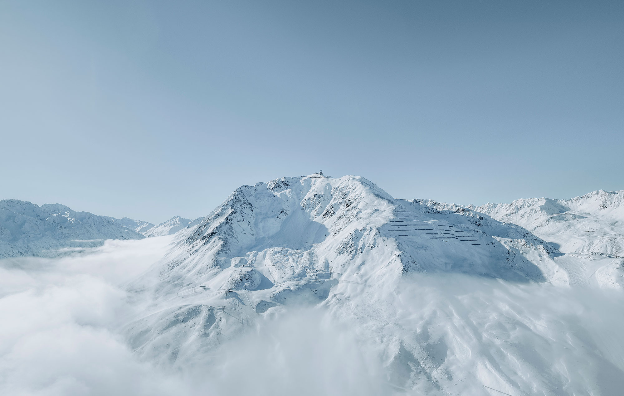 A snow-capped mountain peak rises above the clouds beneath a clear, light blue sky. Distant mountains are in the background.