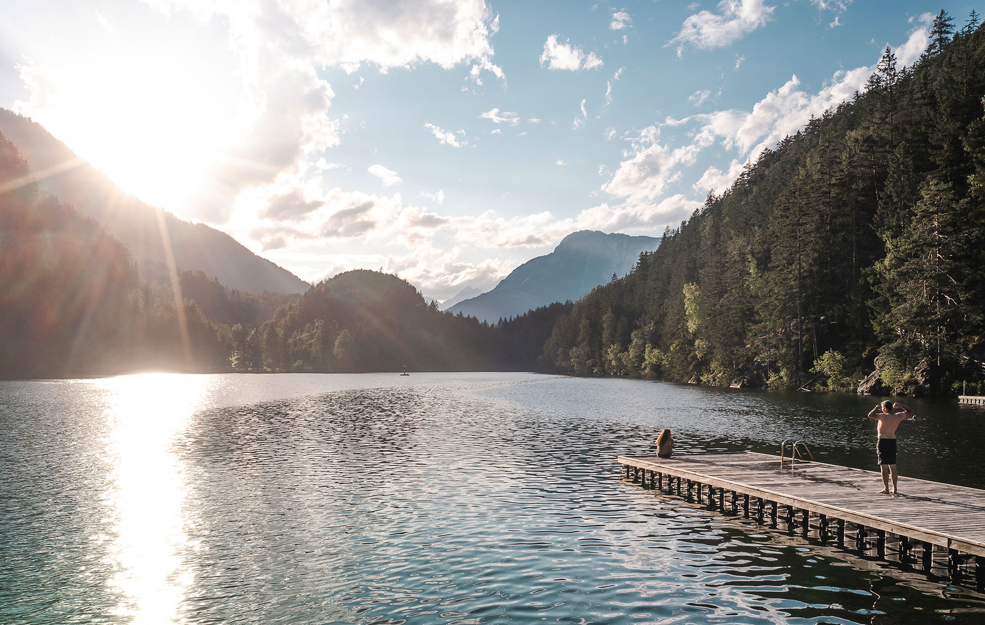 A person stands on a wooden jetty next to a calm lake, surrounded by mountains and trees while sunlight shines through the clouds and reflects on the water.
