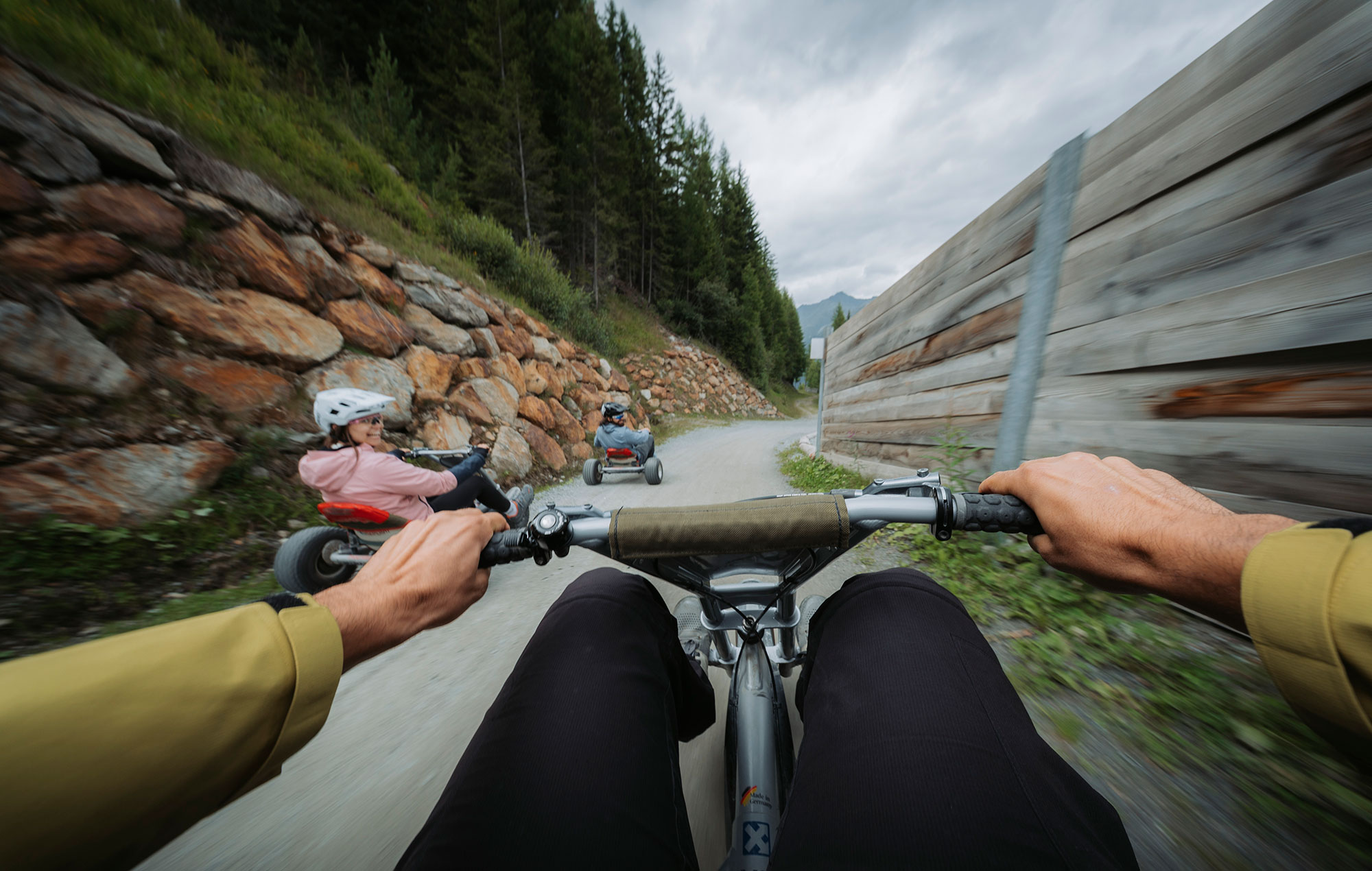 A shot from the perspective of someone riding a tricycle down a winding outdoor path, with two other people riding ahead: all are wearing helmets. Trees and a stone wall line the route. The image conveys a sense of speed and movement.