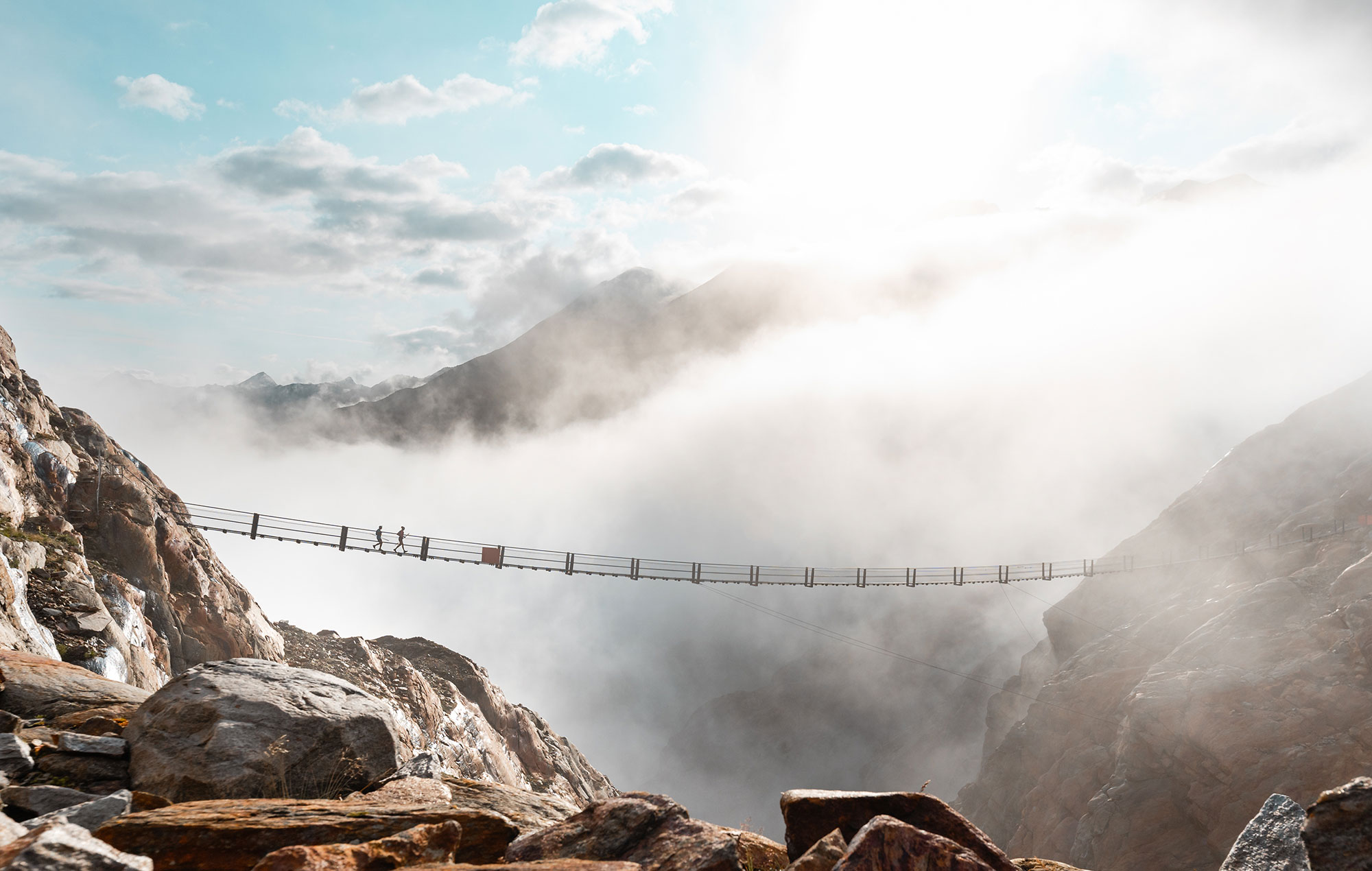 A narrow suspension bridge spans a deep, mist-shrouded mountain gorge, across which several people are walking beneath a bright, cloudy sky. Rugged rocks frame the foreground.
