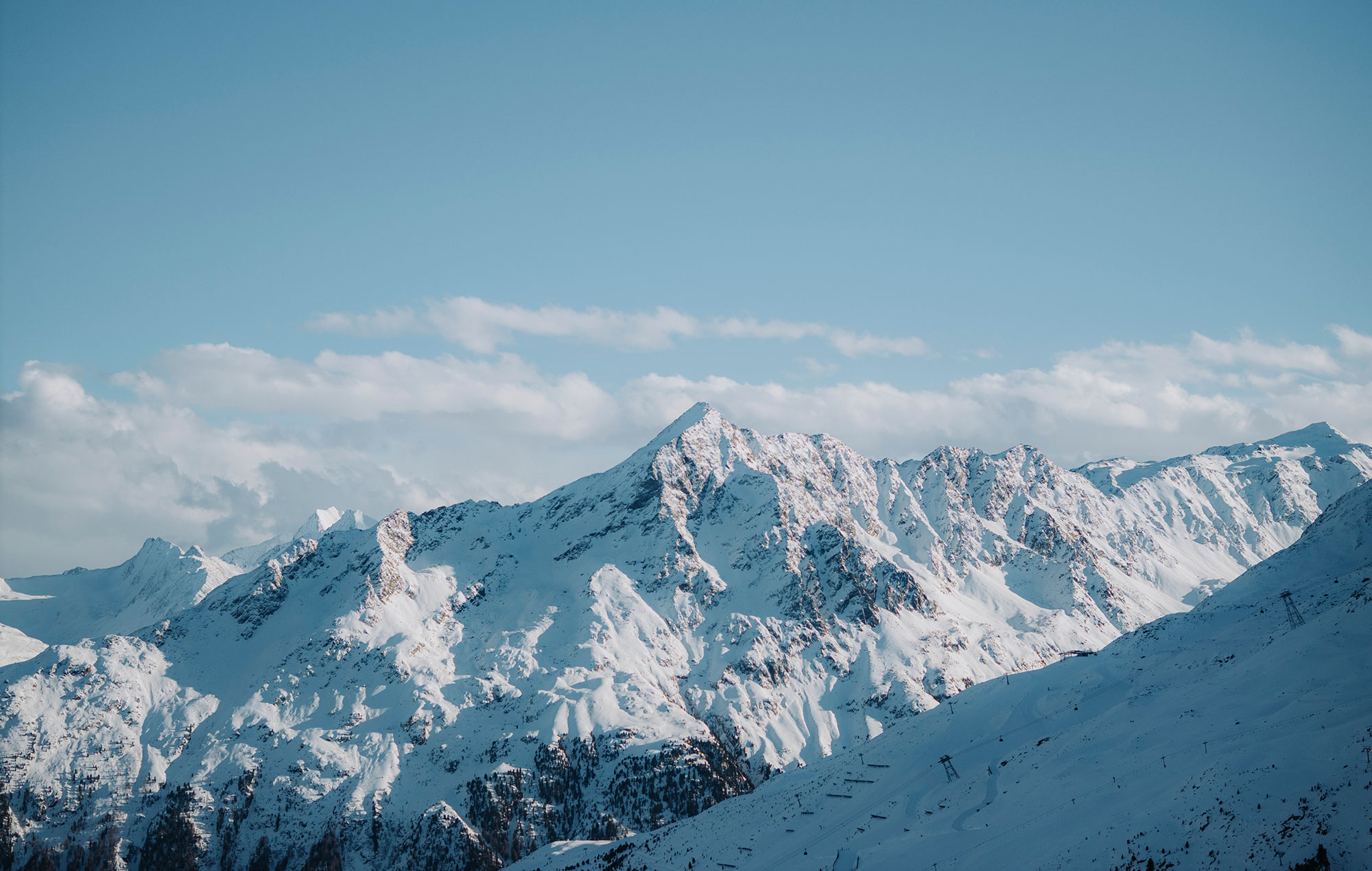 A snow-capped mountain range under a brilliant blue sky with rugged peaks and scattered clouds in the background.