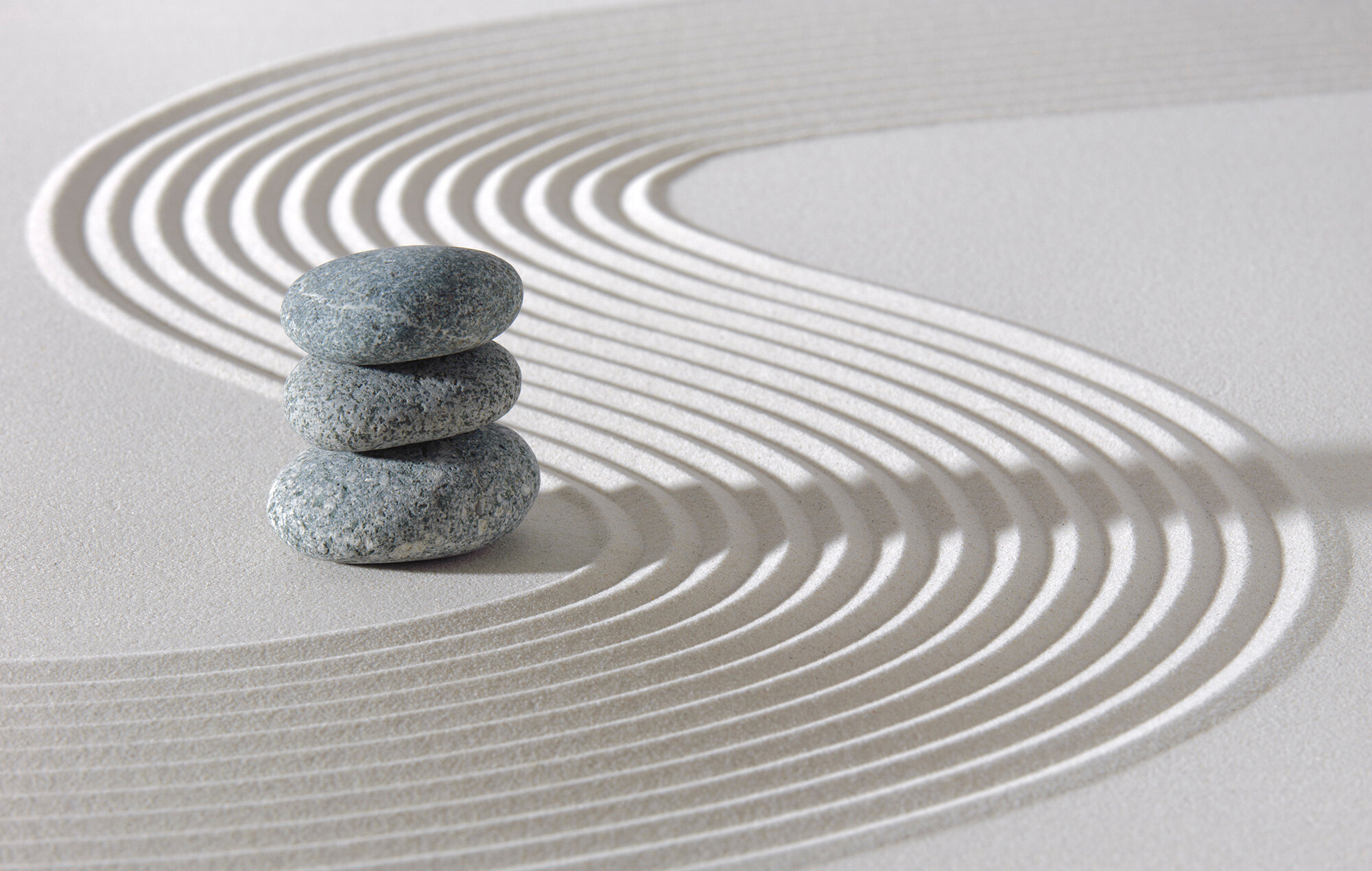 Drei glatte graue Steine, die auf weißem Sand mit geschwungenen, parallelen Linien aufgeschichtet sind. Die Komposition strahlt Ruhe und Minimalismus aus und erinnert an einen japanischen Zen-Garten.