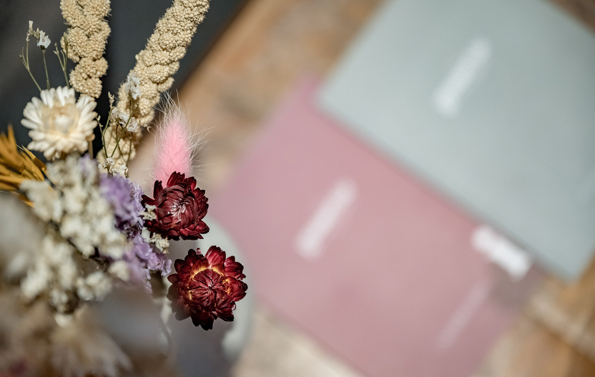 A close-up of a bouquet of dried flowers in the foreground, consisting of red and white blossoms. In the background, blurred pink and green brochures lie on a wooden surface.