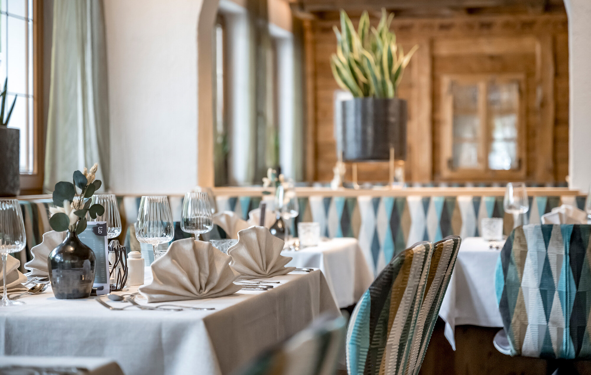 A cozy restaurant with white tablecloths, folded napkins, glassware, patterned chairs, potted plants and large windows through which daylight streams in.
