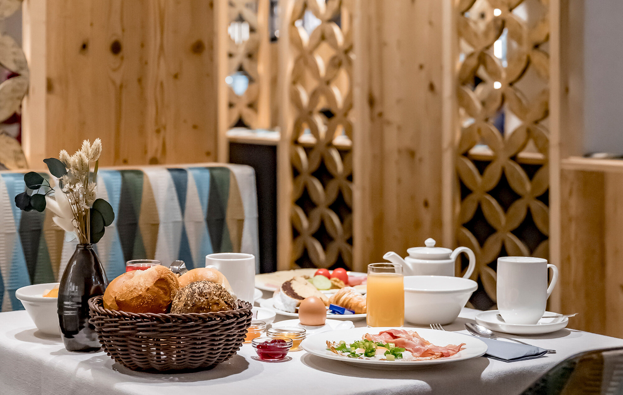 A breakfast table set with bread rolls in a basket, sliced ​​bread, bacon, an egg, orange juice, tea, coffee cups and a black vase with dried flowers, in front of a decorative wooden wall.