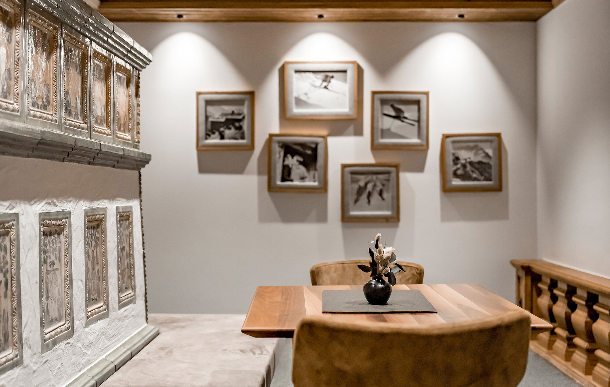A cozy seating area with a wooden table, brown chairs and a decorative vase. Behind it, six framed black-and-white photos hang on a white wall illuminated by spotlights.