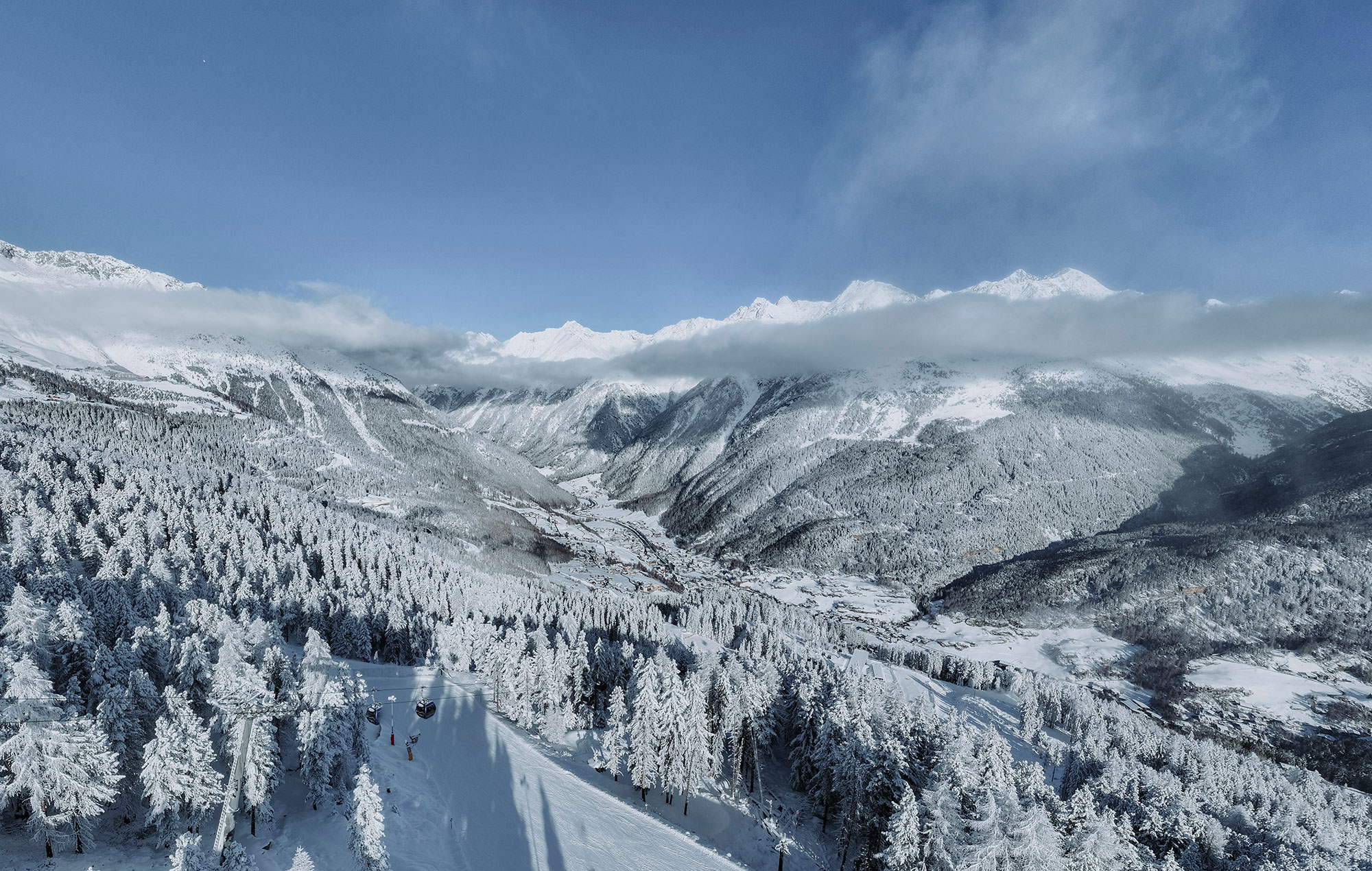 Snow-capped mountains and stone pine trees beneath a blue sky with scattered clouds. A valley runs through the center, flanked by frost-covered forests and hills, creating a peaceful winter landscape.