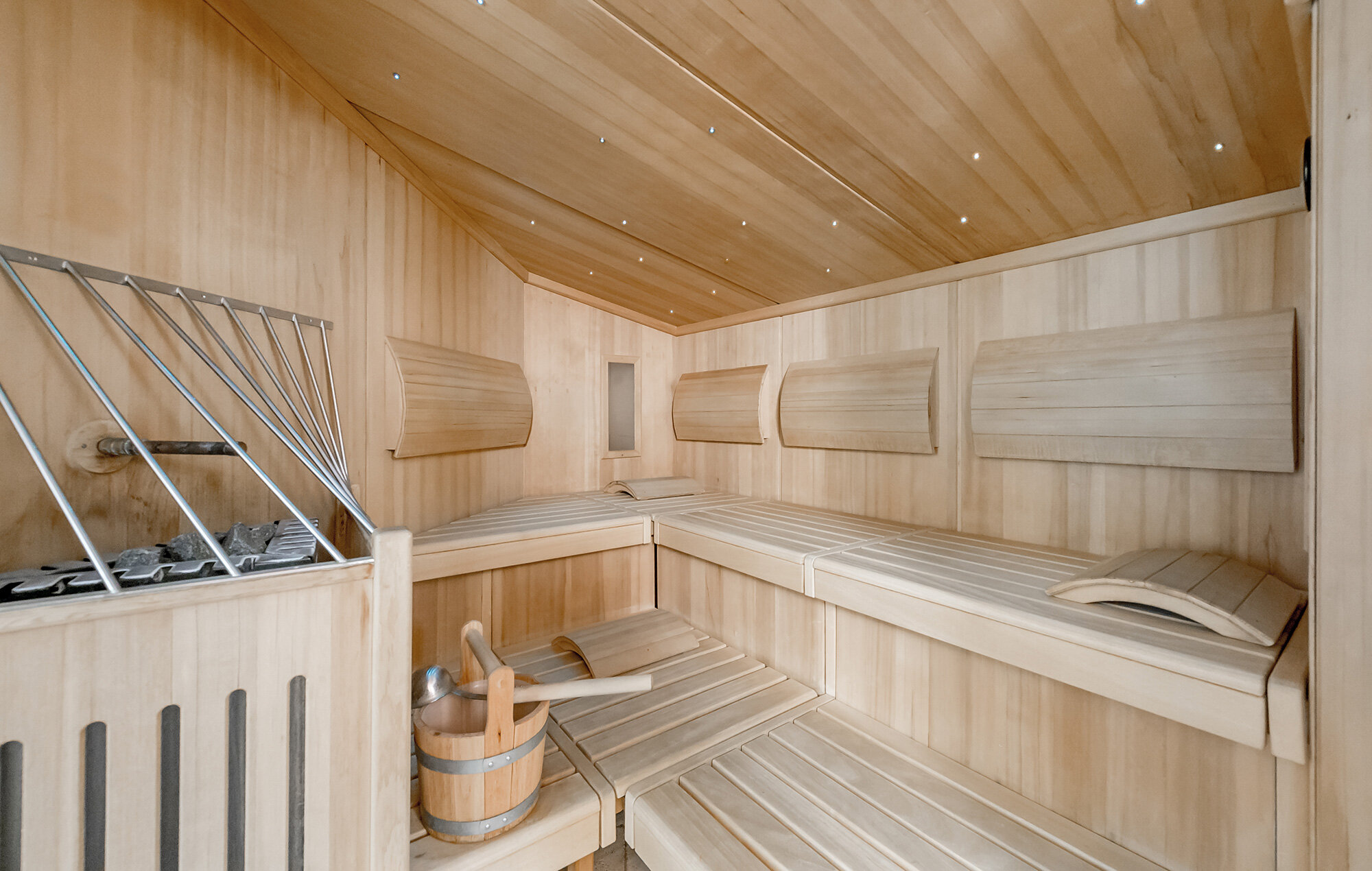 A modern wooden sauna featuring benches, a heater, a bucket with a ladle and headrests. The walls and ceiling are paneled in light-colored wood, creating a warm and inviting atmosphere.